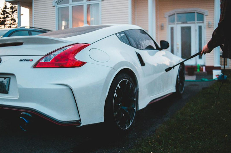 A person rinsing a white sports car with a pressure washer in a driveway